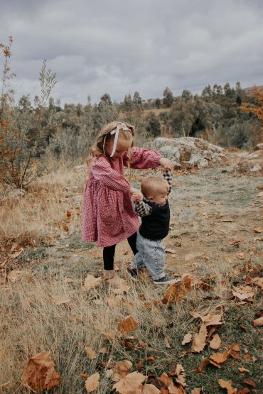 Older girl helps toddler walk outdoors in autumn setting.
