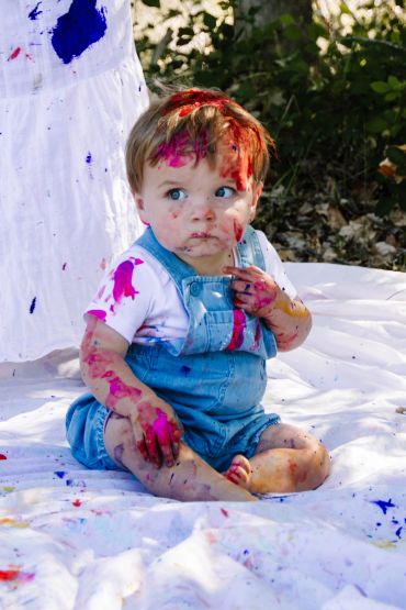 A baby covered in colorful paint sitting on a white blanket.