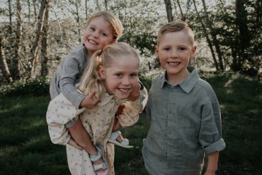 Three smiling children outdoors, with one girl giving a piggyback ride.