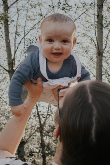 Adult lifting smiling baby outdoors.