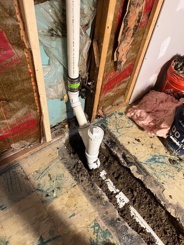 White plumbing pipe beside an open floor trench and exposed wall framing.