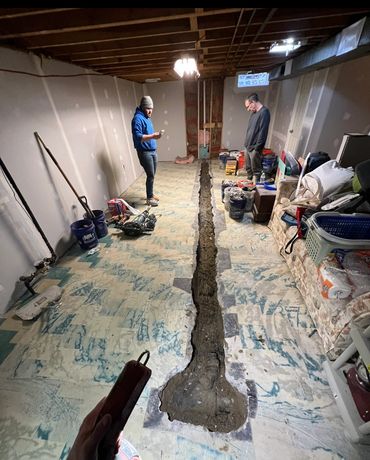 Two people standing beside an open trench in a basement floor.