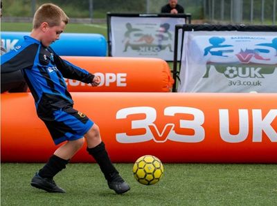 A young boy in blue and black soccer kit kicks a yellow ball on a field with an inflatable 3v3 UK sign.