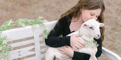 A girl in a black dress hugging a white lamb on a bench decorated with greenery.