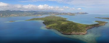Aerial view of a lush island surrounded by clear blue waters under a partly cloudy sky.