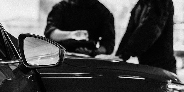 Two people conversing near a car's side mirror in black and white.