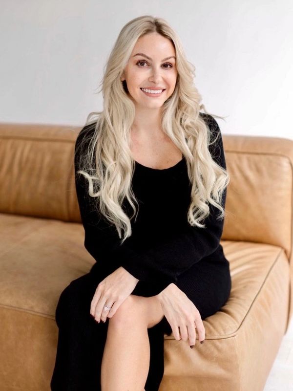 Smiling woman with long blonde hair in a black dress sitting on a brown leather couch.
