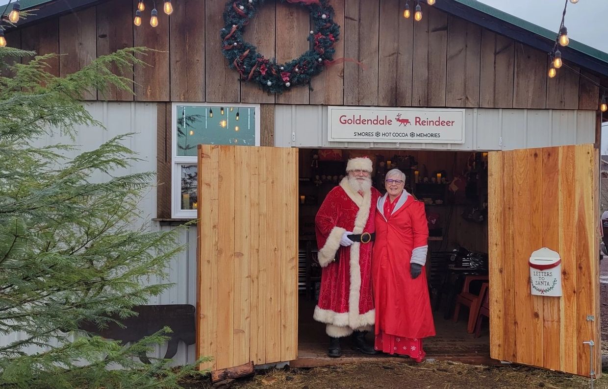 Santa and a woman in red coats stand at a festive doorway with holiday decorations.