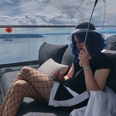 Person with blue hair sitting on a couch by the water, holding a transparent umbrella.