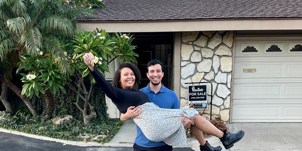 Happy couple celebrating in front of their new home with a for sale sign.