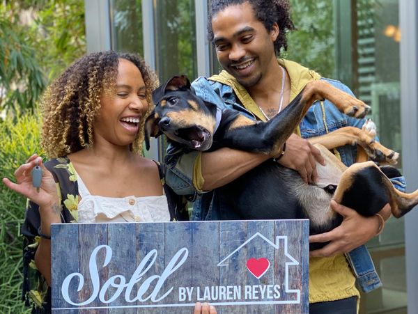 Happy couple holds a dog and a sold sign in front of their new home.