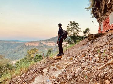 A person stands on a rocky hillside overlooking a scenic valley at sunset.