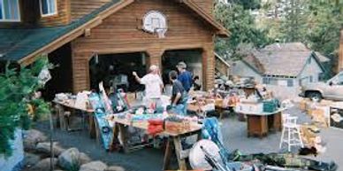 People browsing items at a garage sale outside a wooden house.