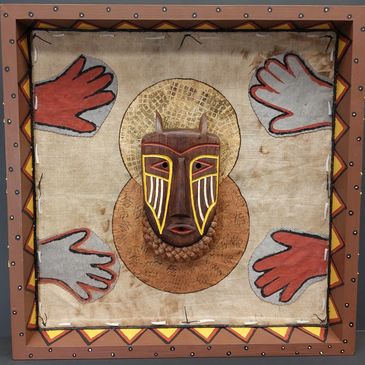 Framed tribal mask with painted hands and geometric border on fabric background.