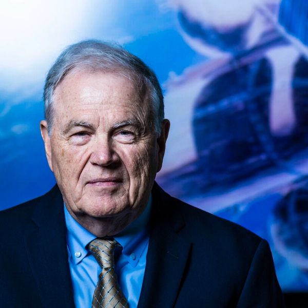 Dr Tom Edwards, an older white man in a suit and tie stands in front of a blue-toned background