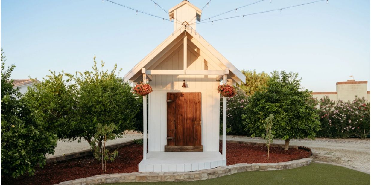 Small white wooden playhouse with a brown door, surrounded by greenery and flowers.
