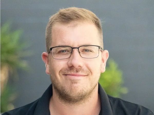 Smiling man in black polo shirt and glasses against a blurred outdoor background.