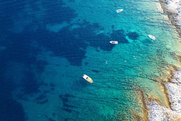 Aerial view of boats and swimmers in clear turquoise sea near rocky shore.