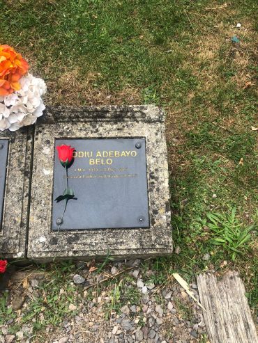 Grave marker with a red rose and flowers on grass and gravel.