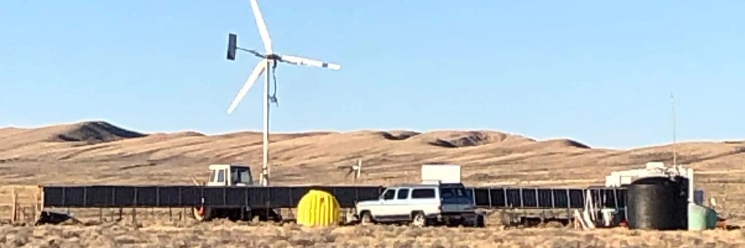 Photograph of 100 Acres Ranch. A bunch of solar panels, a windmill, a tractor. Mountains behind.