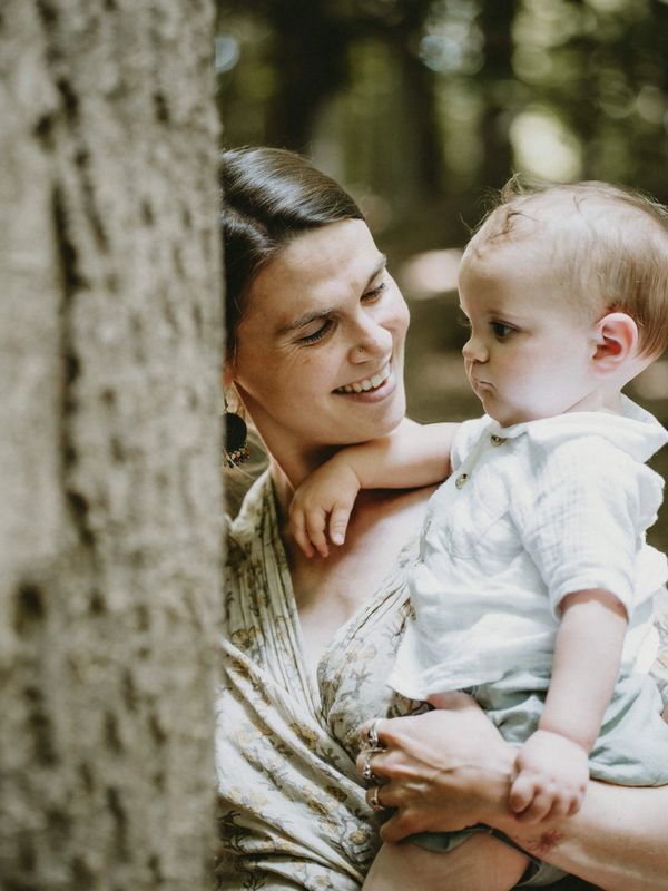 A Bath Doula gazes lovingly towards her child in the woods next to a tree