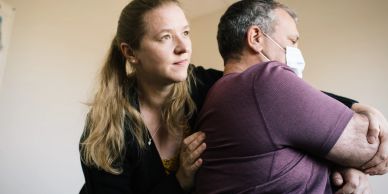 A woman comforts a man wearing a face mask indoors.