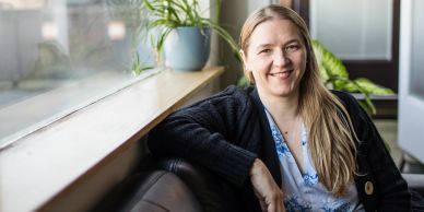 Smiling woman sitting on a couch by a window with plants.