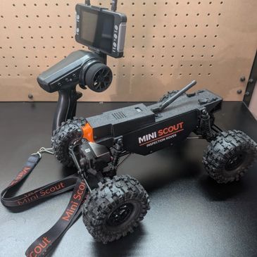 A Mini Scout inspection rover with a remote control and monitor on a black surface.