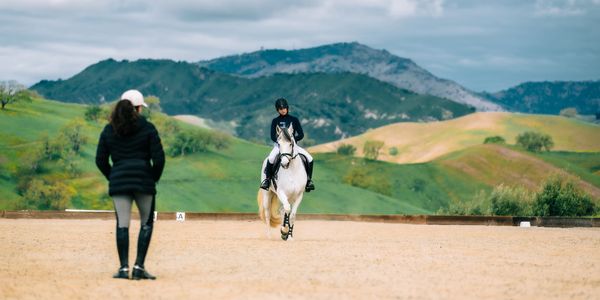 Person riding a horse with an observer in a scenic outdoor arena.