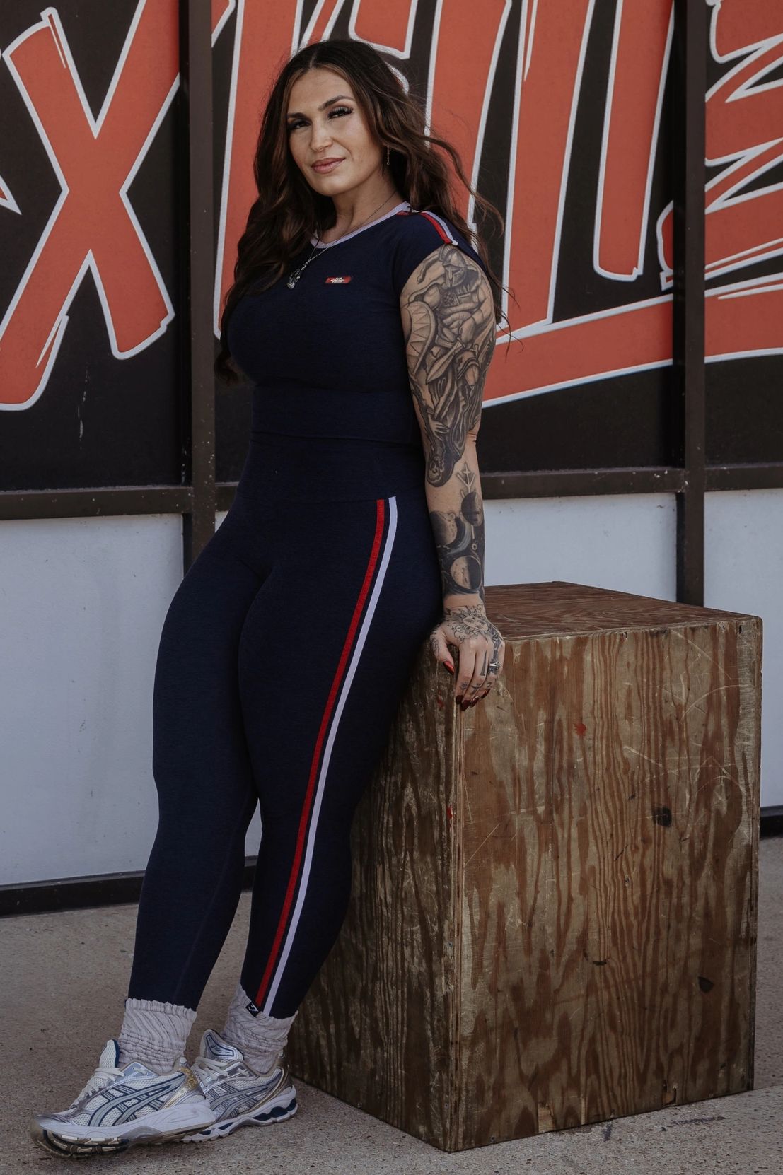 Woman with tattoos posing by a 'PROJECT SIXTILZ' wall.
