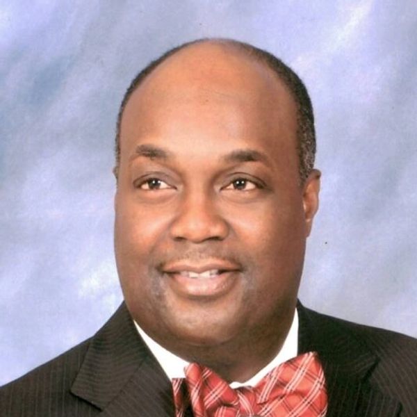 A man in a suit with a red bow tie smiling against a light blue background.