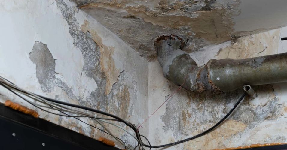 Rusty pipes and peeling paint on a deteriorating ceiling corner with exposed wires.