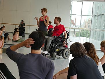 Workshop: Luke, who dances using a wheelchair & Kathleen coteaching students in a bright studio.