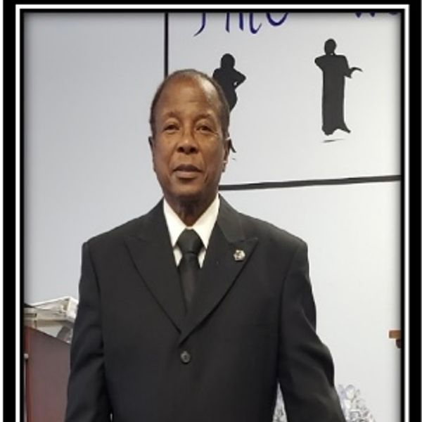 A man in a black suit and tie posing indoors.
