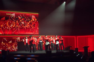 Performers in red jackets on stage with vibrant red background and lighting.