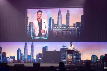 Speaker presenting with city skyline and Petronas Towers backdrop on stage.