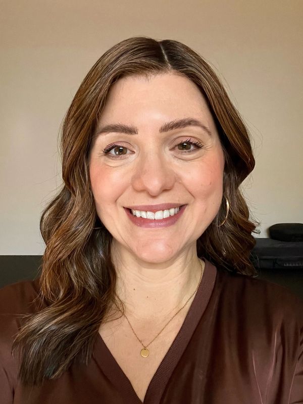 Smiling woman with wavy brown hair wearing a brown top and gold necklace.