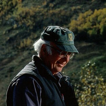 Elderly man smiling outdoors wearing a cap and glasses.