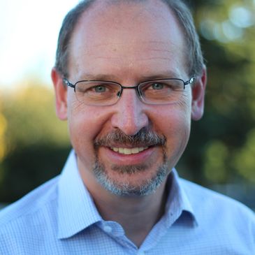 Smiling middle-aged man wearing glasses and a light blue shirt outdoors.