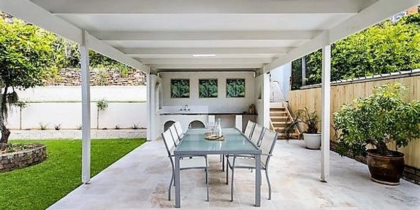 Table with 8 chairs on paved area under pergola surrounded by garden with stairs to the right side 