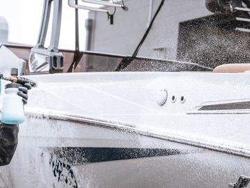 Person washing a white boat with a spray bottle and water hose.