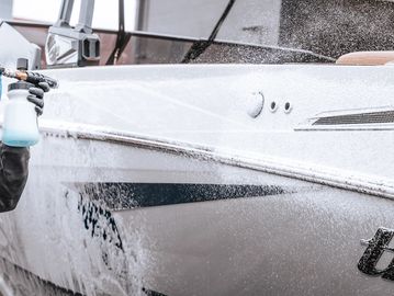 Person washing a white boat with a spray bottle and water hose.