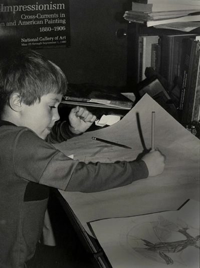 A young boy focused on drawing at a cluttered desk.