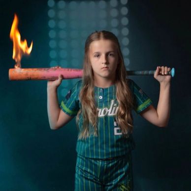 Young girl in baseball uniform holding a flaming bat with a serious expression.