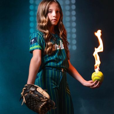 Young girl in baseball uniform holding a flaming softball and glove.