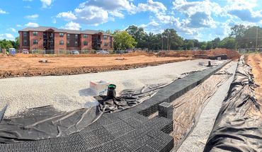 Construction site with gravel foundation and drainage system, near apartments under a sunny sky.