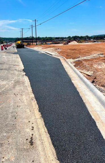 Freshly paved road section in construction area with curb, utility poles, and dirt surroundings.