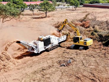 Excavator loading soil into a large grinder machine on a cleared construction site.