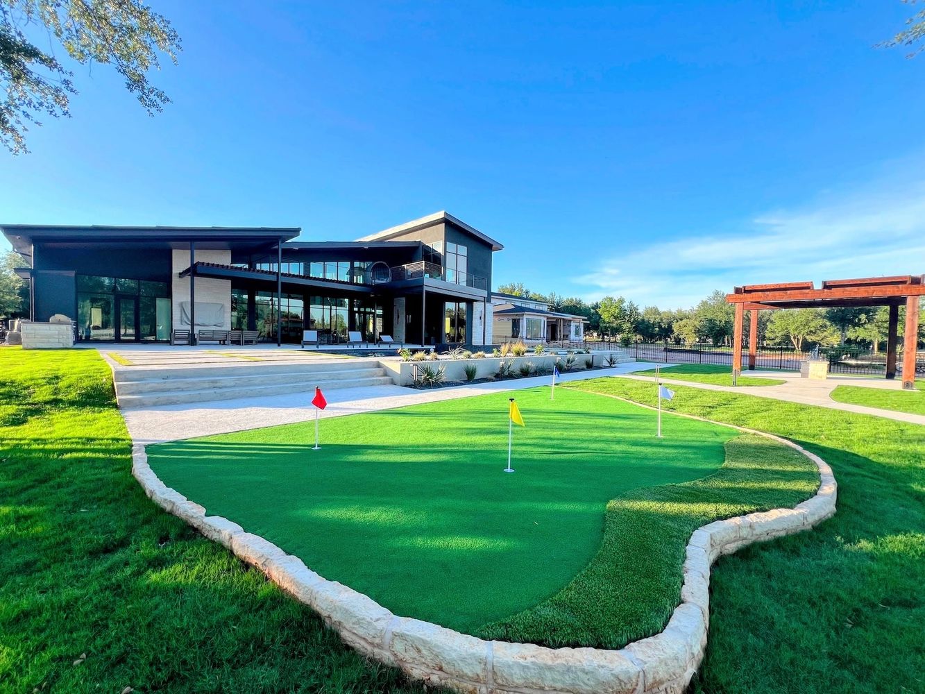 Modern glass building with putting green and pergola on landscaped property.