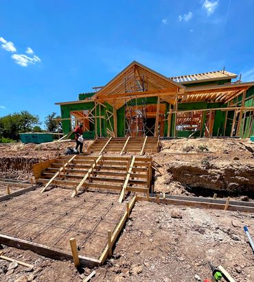 Framed house under construction with front steps being prepared, worker on site under clear sky.
